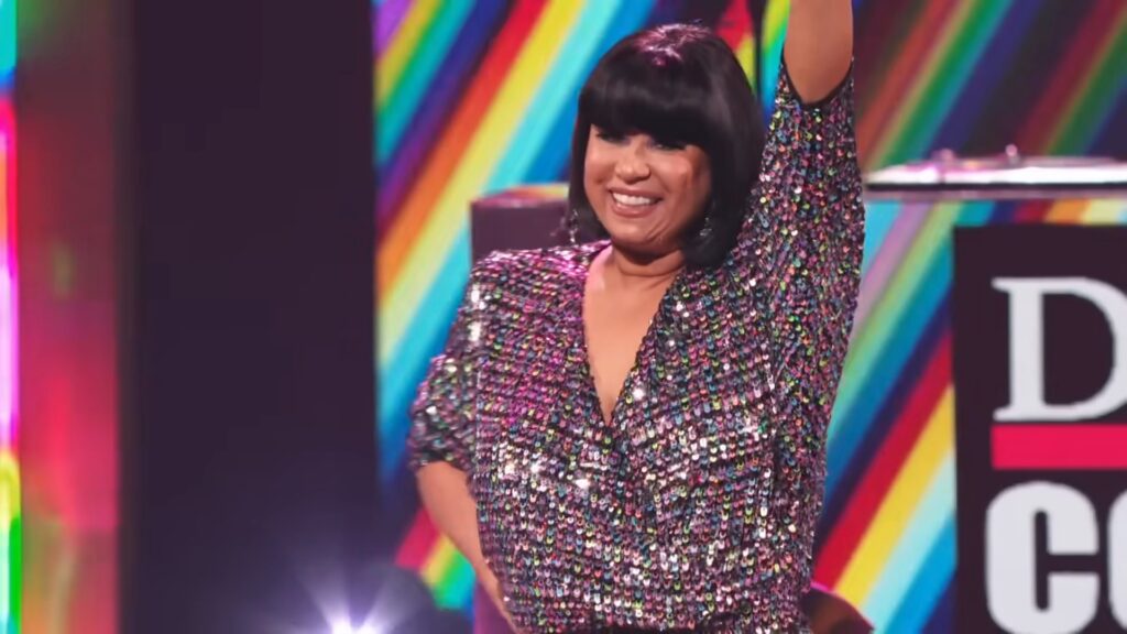 CeCe Peniston performing onstage in a multicolored sequin outfit, smiling with one arm raised against a vibrant rainbow backdrop.