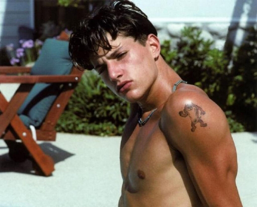 Young Simon Rex outdoors in the early 1990s, shirtless with a Taz tattoo on his upper arm.