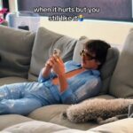 Benson Boone at home wearing his Grammy performance Blue Jump Suit laying on a oversized couch while scrolling on his phone.