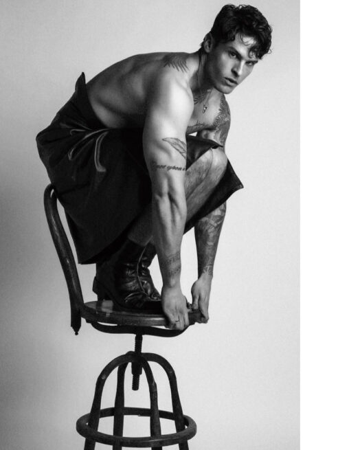 Black-and-white portrait of Baptiste Giabiconi crouching shirtless on a metal stool in dark draped clothing and boots