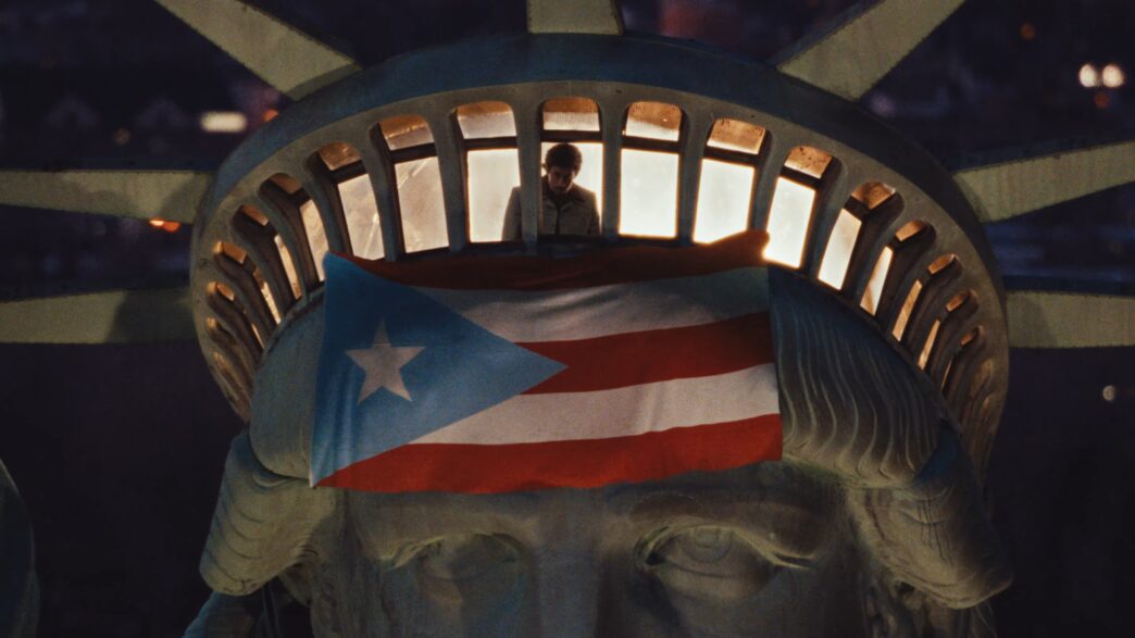 The image shows the head of the Statue of Liberty at night, with a person standing inside the crown's observation deck. A large Puerto Rican flag is draped over the statue's forehead, partially covering its eyes.