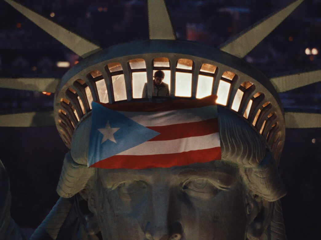 The image shows the head of the Statue of Liberty at night, with a person standing inside the crown's observation deck. A large Puerto Rican flag is draped over the statue's forehead, partially covering its eyes.
