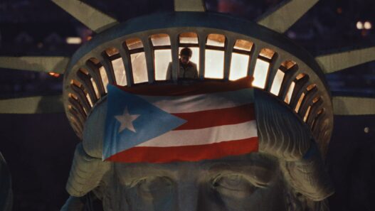 The image shows the head of the Statue of Liberty at night, with a person standing inside the crown's observation deck. A large Puerto Rican flag is draped over the statue's forehead, partially covering its eyes.