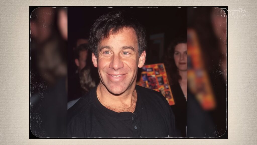 Alan Menken smiling at an event, wearing a black shirt, with a blurred crowd in the background