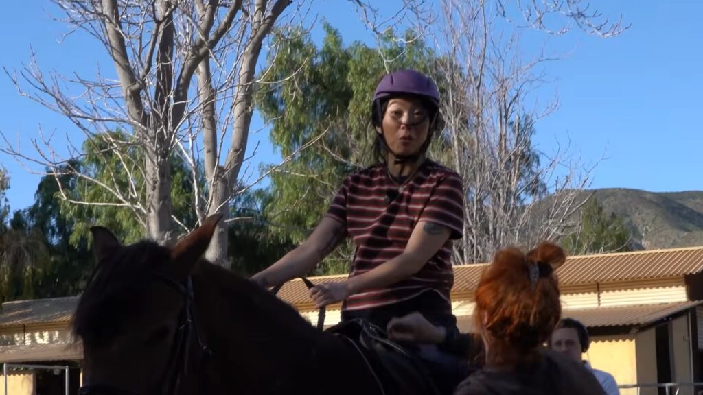 Awkwafina sitting on a horse during a riding lesson, guided by an instructor outdoors