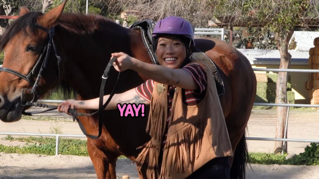 Awkwafina smiling and pointing while standing beside a horse during her Lunar New Year riding lesson