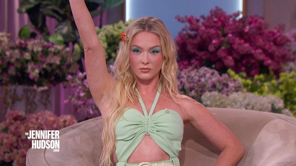 Zara Larsson raising her arm while seated on The Jennifer Hudson Show set, wearing a light green halter look surrounded by vibrant floral décor