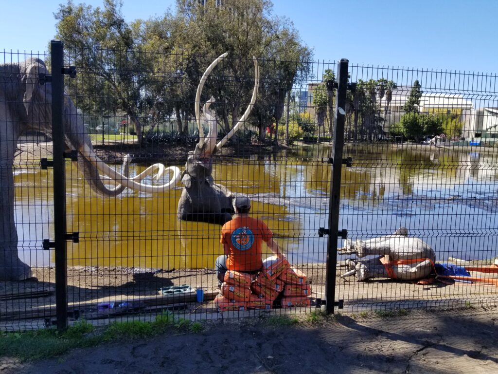 A worker sits near sandbags during mammoth sculpture restoration at the La Brea Tar Pits in Los Angeles, with the tar pond and museum grounds visible behind a fence.