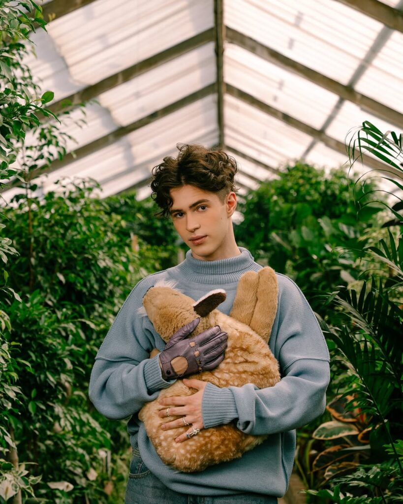 Mikhail Shaidorov stands in a sunlit greenhouse, wearing a light blue turtleneck and holding a plush animal among vibrant green plants.