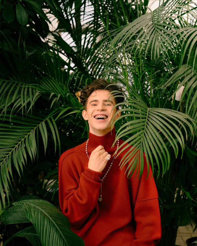 A joyful Mikhail Shaidorov poses in a greenhouse of vibrant greenery, wearing a red turtleneck.