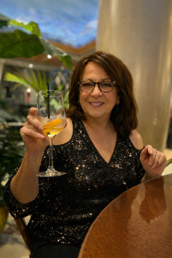 Patti Masi of Vera in a softly enhanced portrait wearing a dark elegant diva-style top, subtle glamour makeup and gentle soft focus lighting while raising a cocktail glass.