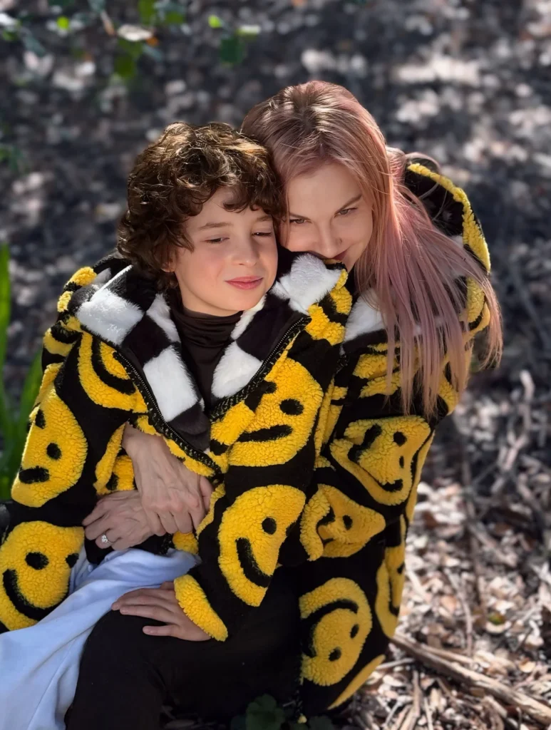 Max Alexander sits with his mother in coordinated smiley‑pattern jackets among fallen leaves.