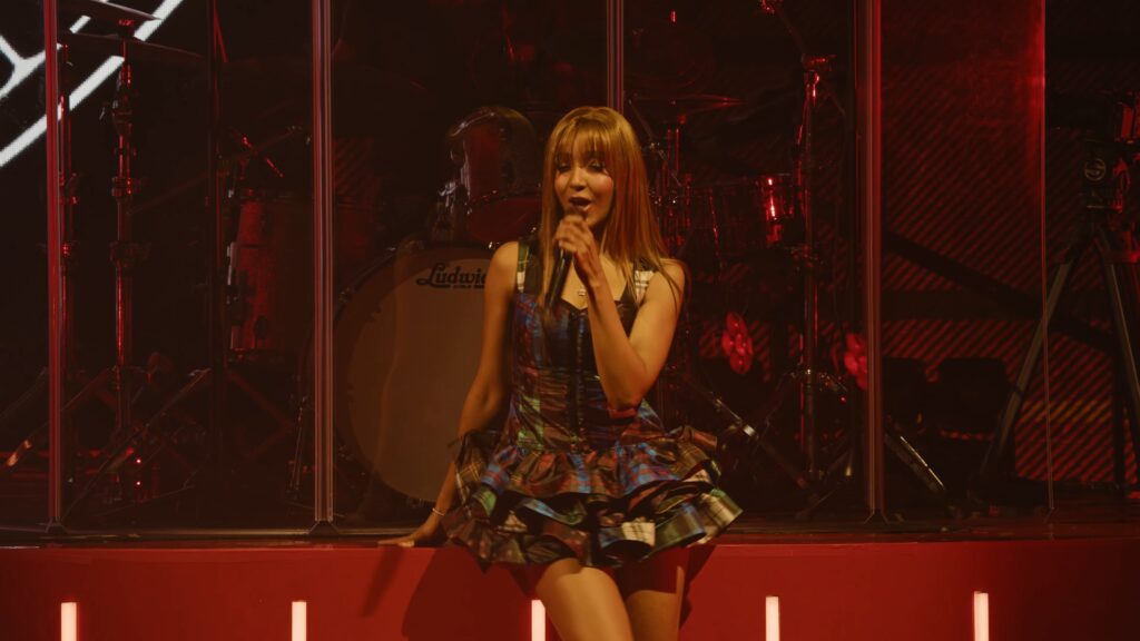 PinkPantheress performing live at the Wiltern in a patterned dress, singing into the mic under red stage lighting.