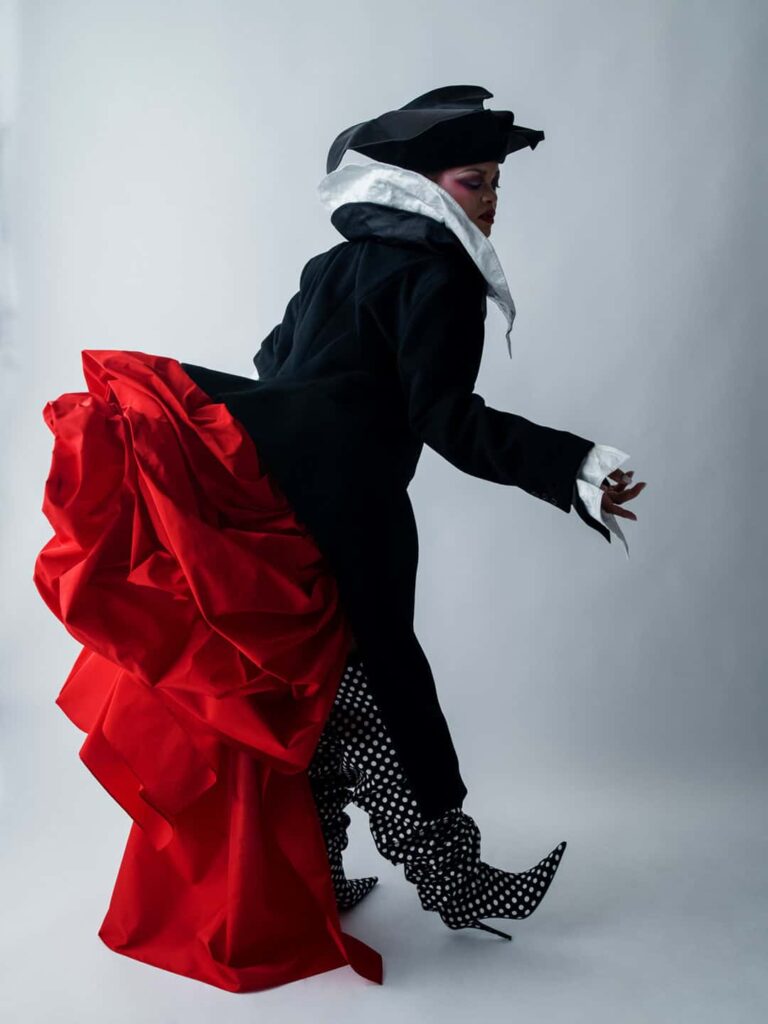 Person in a black coat with a white scarf, a bright red fabric train sweeping behind, and polka-dot boots in mid-stride in a studio.