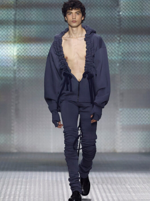 Look from AAA STUDIO’s “Señora, Suélteme El Brazo” runway collection.. Male model on a runway wearing a navy blue oversized hoodie with ruffled trim, unzipped at the chest, and matching joggers with drawstrings.