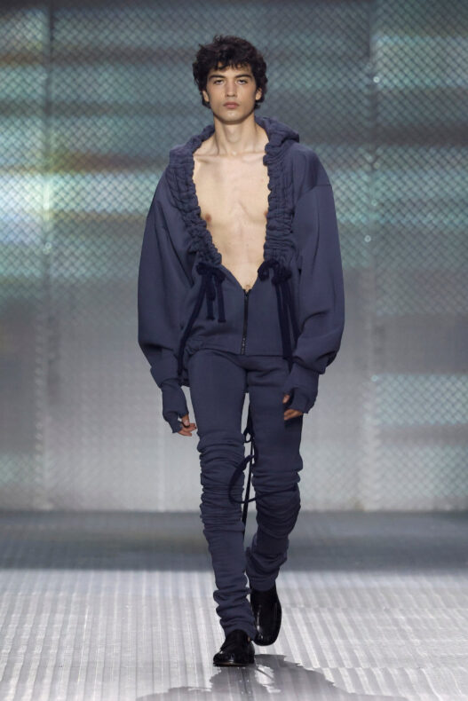 Look from AAA STUDIO’s “Señora, Suélteme El Brazo” runway collection.. Male model on a runway wearing a navy blue oversized hoodie with ruffled trim, unzipped at the chest, and matching joggers with drawstrings.