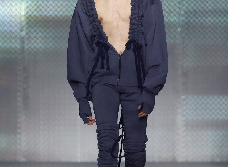 Look from AAA STUDIO’s “Señora, Suélteme El Brazo” runway collection.. Male model on a runway wearing a navy blue oversized hoodie with ruffled trim, unzipped at the chest, and matching joggers with drawstrings.