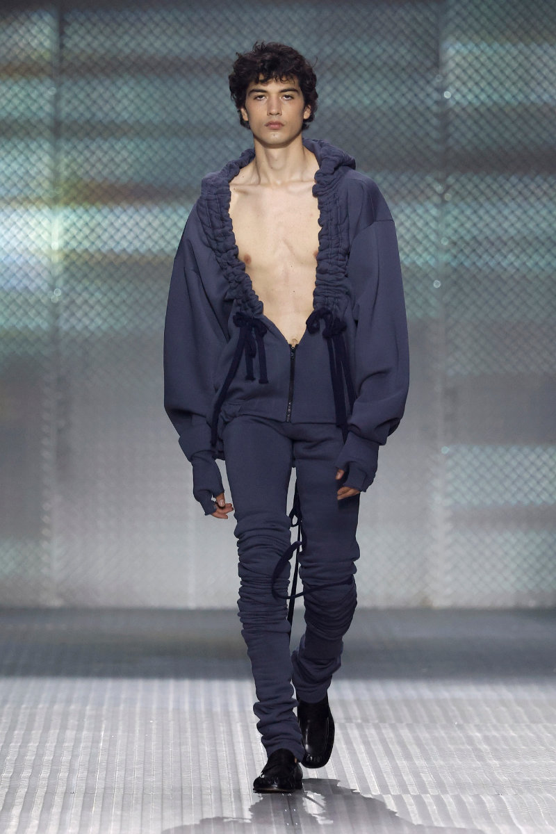Look from AAA STUDIO’s “Señora, Suélteme El Brazo” runway collection.. Male model on a runway wearing a navy blue oversized hoodie with ruffled trim, unzipped at the chest, and matching joggers with drawstrings.