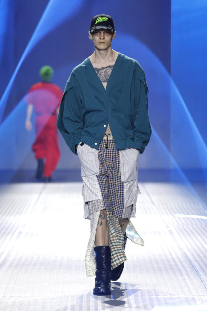 Model walks the runway in a teal oversized jacket over layered plaid skirts, black boots, and a black cap, against a blue stage backdrop.