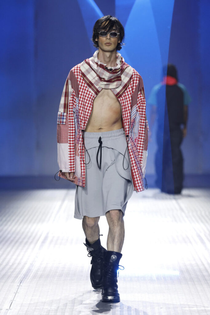 Male model walking a fashion show in a red plaid jacket draped over shoulders, chest exposed, gray drawstring shorts, black combat boots, and sunglasses against a blue-lit runway backdrop.