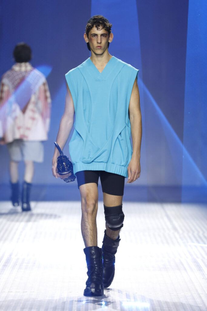 Male model walks the runway in a bright turquoise sleeveless top, black shorts, knee brace, and tall black boots on a lit catwalk.