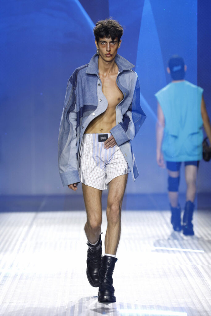 Male model on a runway in an oversized blue denim jacket, bare chest, white striped shorts, and black boots.