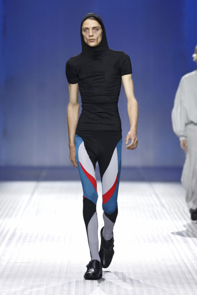 Runway model wearing a black hooded, ruched top with bold color-block leggings in blue, red, gray as she walks the catwalk. Bruises visible on her face.