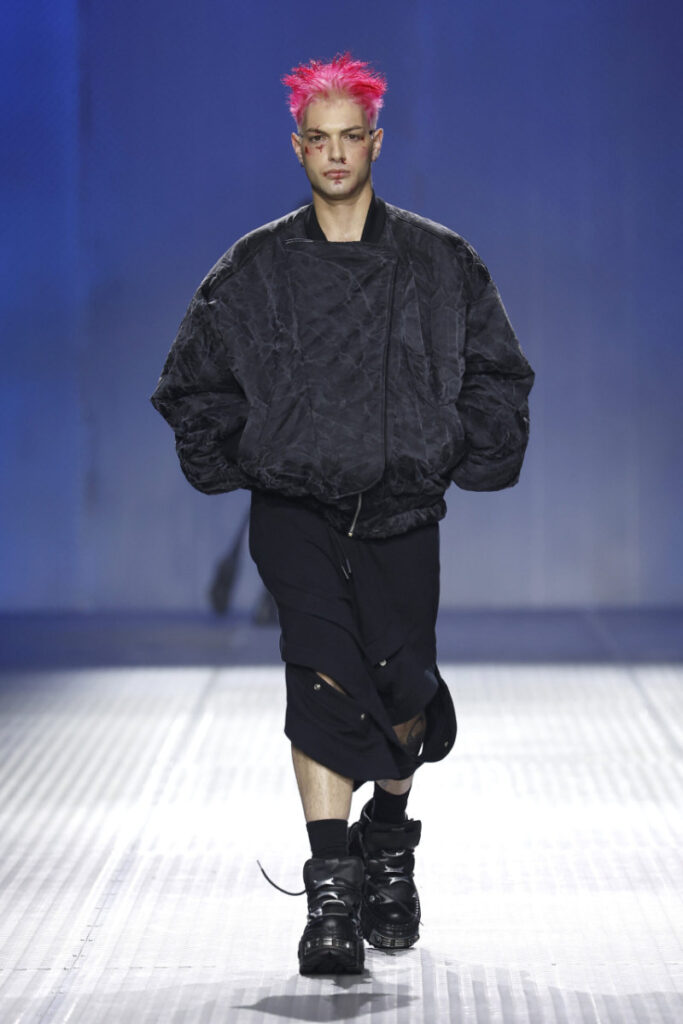 Male model with bright pink spiky hair walks the runway in a oversized black bomber jacket and cropped black pants with chunky boots against a blue backdrop.