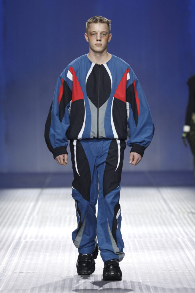 Male model walking a fashion runway in a blue, black, red, and gray color-blocked athletic tracksuit with puffed sleeves and matching pants; black chunky shoes.