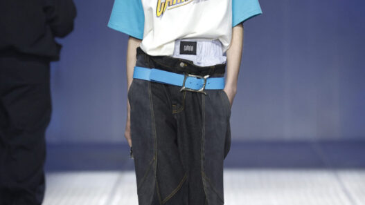 Runway model walking the catwalk in a white-and-teal Carieri T-shirt, oversized dark jeans, and a bright blue belt with black boots.