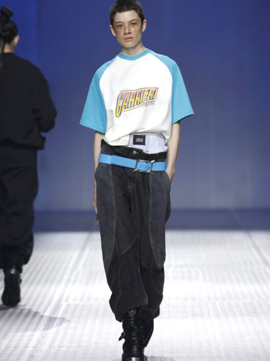 Runway model walking the catwalk in a white-and-teal Carieri T-shirt, oversized dark jeans, and a bright blue belt with black boots.