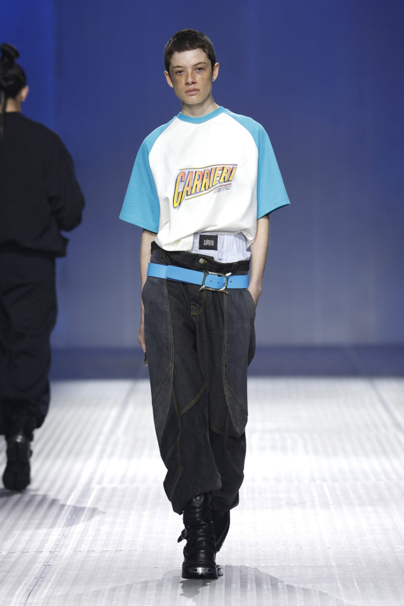 Runway model walking the catwalk in a white-and-teal Carieri T-shirt, oversized dark jeans, and a bright blue belt with black boots.