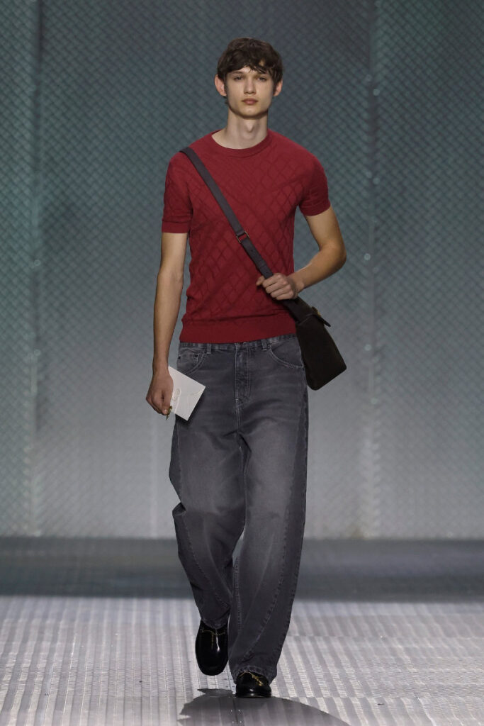 Male model on a runway wearing a red knit T-shirt, loose gray jeans, black shoes, and a crossbody bag while holding a white card
