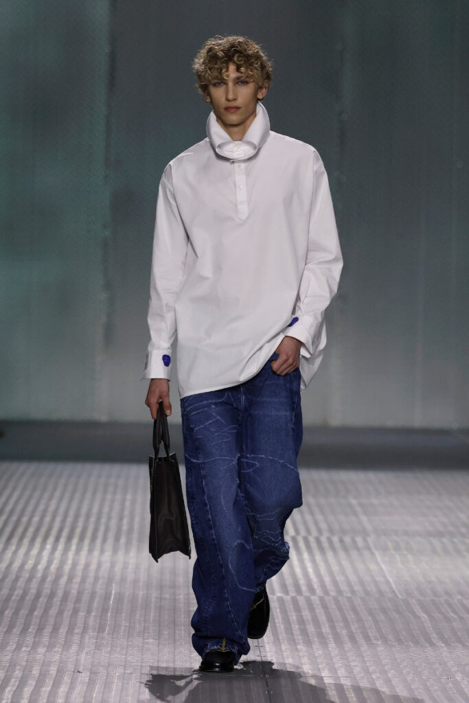 Male model walking a runway in an oversized white shirt and baggy blue jeans, carrying a small black bag.