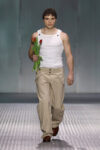Male model on a fashion runway walks toward camera, wearing a white sleeveless top and beige pants, holding red tulips by his side.