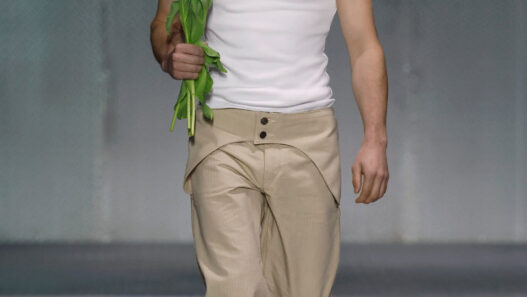 Male model on a fashion runway walks toward camera, wearing a white sleeveless top and beige pants, holding red tulips by his side.