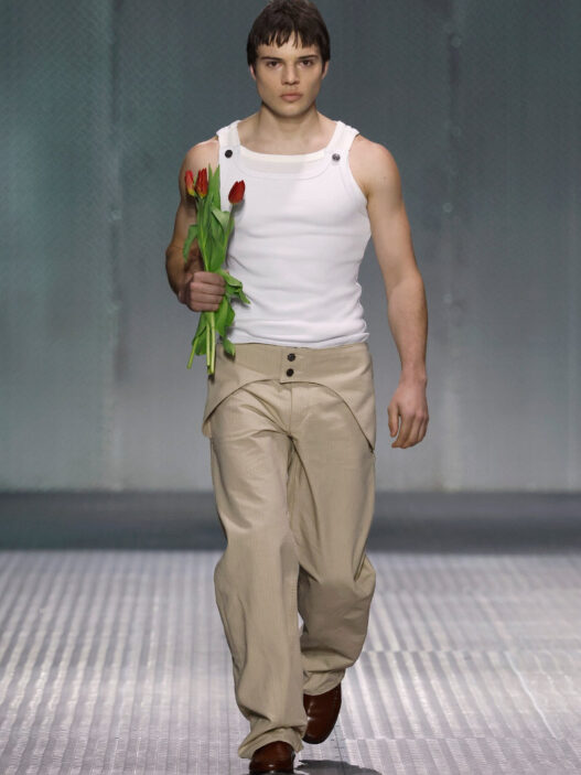Male model on a fashion runway walks toward camera, wearing a white sleeveless top and beige pants, holding red tulips by his side.