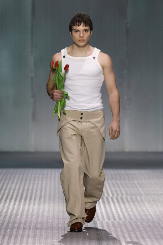 Male model on a fashion runway walks toward camera, wearing a white sleeveless top and beige pants, holding red tulips by his side.