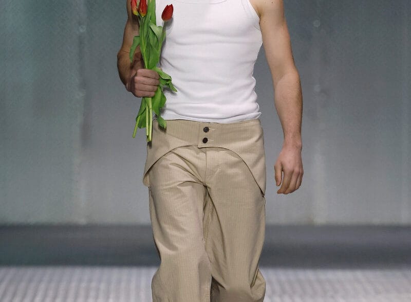 Male model on a fashion runway walks toward camera, wearing a white sleeveless top and beige pants, holding red tulips by his side.