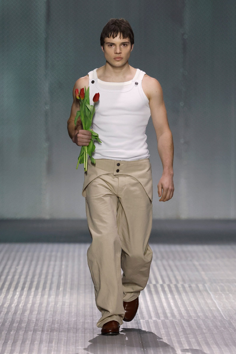 Male model on a fashion runway walks toward camera, wearing a white sleeveless top and beige pants, holding red tulips by his side.