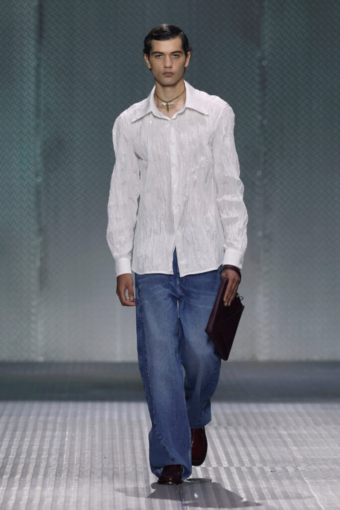 Male model on a runway wearing a white textured shirt, loose blue jeans, dark burgundy shoes, and carrying a burgundy clutch.
