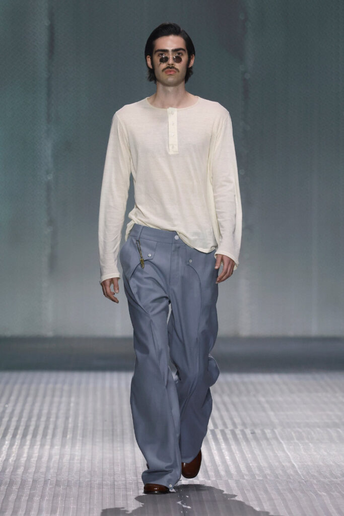 Male fashion model walks the runway in a cream long-sleeve henley and light blue wide-leg pants, with round sunglasses and a mustache.