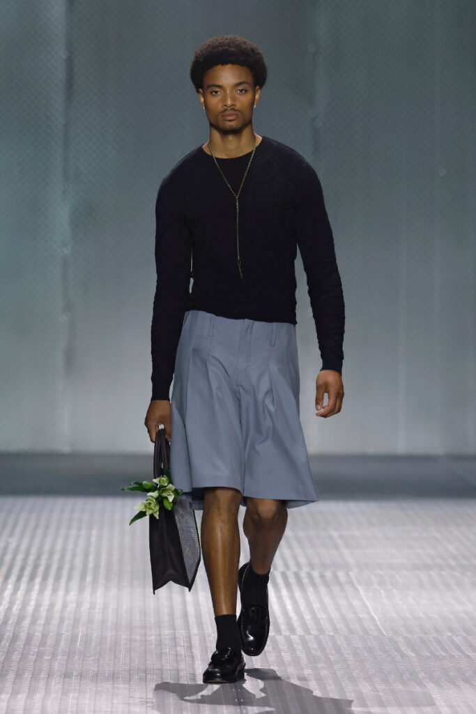 Male model walks a runway in a black long-sleeve top, light gray knee-length shorts, black loafers, and a small black tote with white flowers tucked inside.