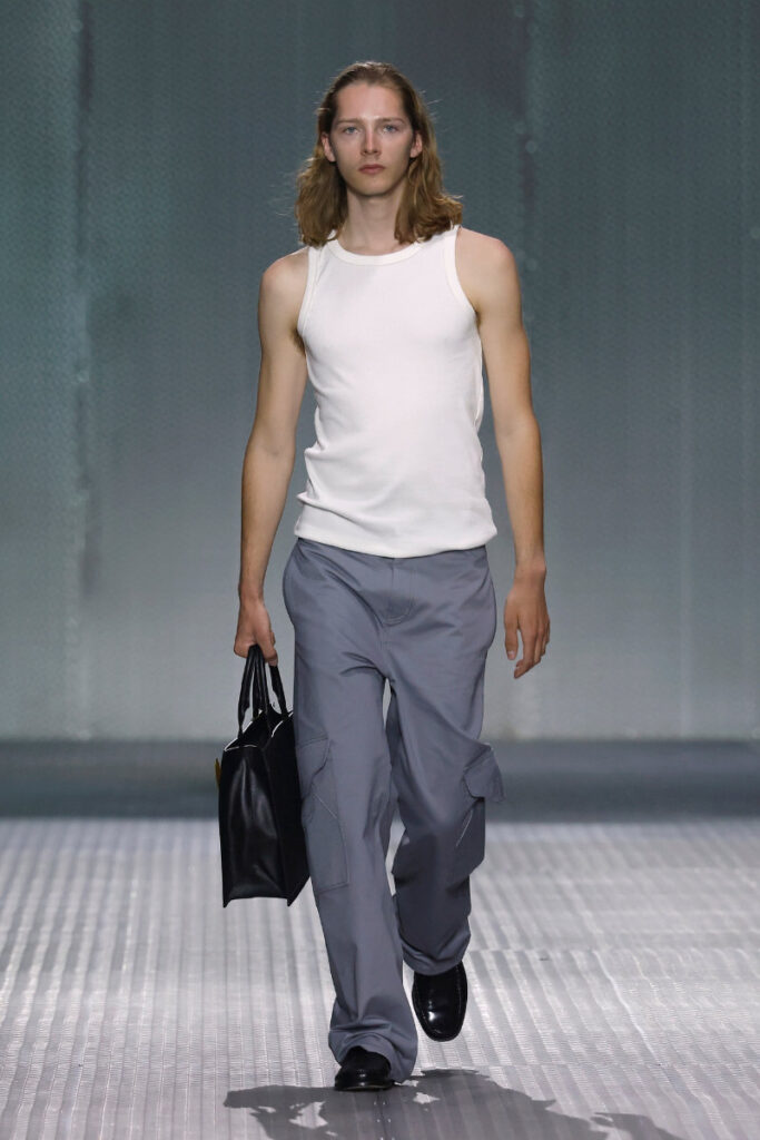Male model walks a runway in a white sleeveless top, gray cargo pants, and black shoes, carrying a black bag.