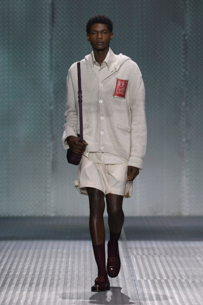Male model walking a fashion runway in a cream cardigan with a red patch, beige shirt, matching shorts, burgundy socks and loafers, carrying a burgundy shoulder bag.