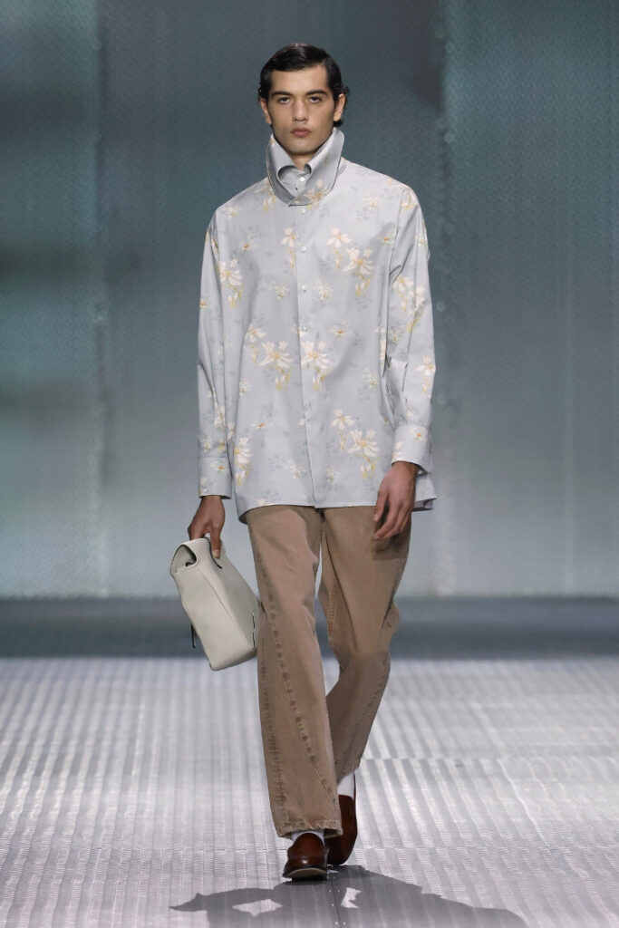 Male model walks a runway in a light blue floral shirt, beige pants, brown shoes, and carries a white clutch.