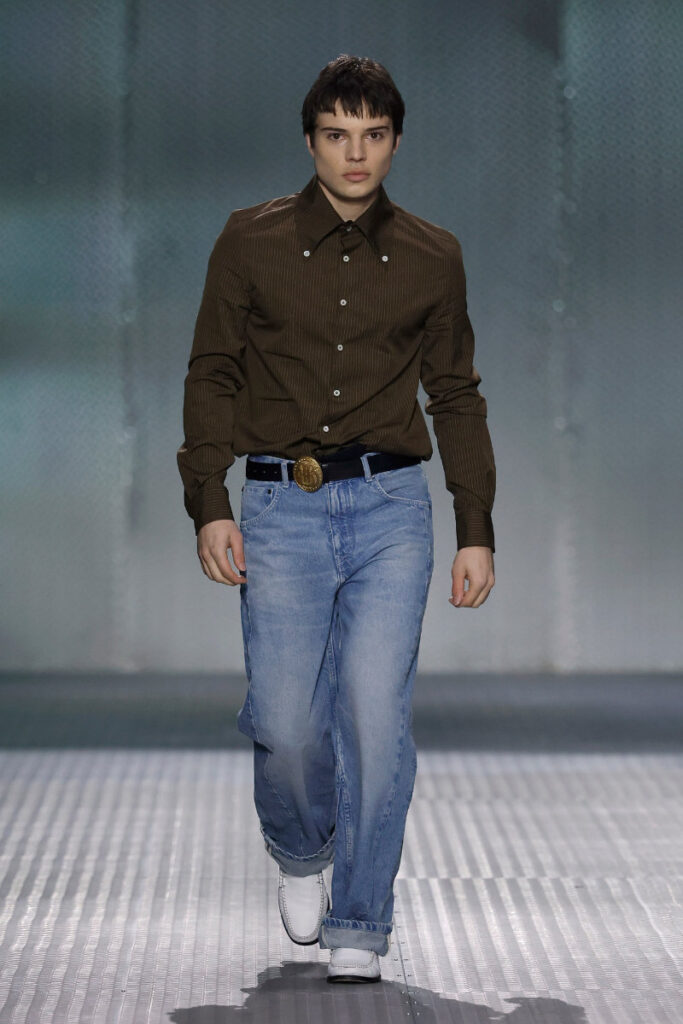 Male model walking a fashion runway in a brown button-down shirt, blue jeans, and a large belt buckle.