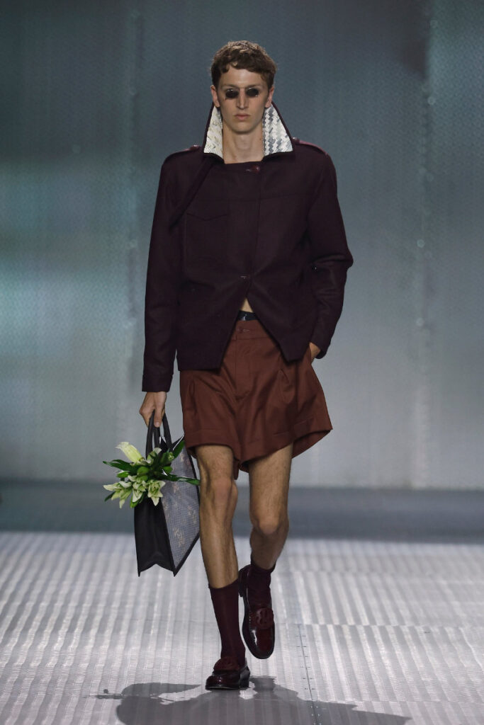 Male model walks a runway in a burgundy jacket with a patterned collar, brown shorts, burgundy socks and loafers, wearing sunglasses and carrying a black tote with flowers.