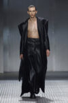 Male model walking runway in a long black leather coat open to reveal bare chest and black wide-leg pants, glossy slicked-back hair.