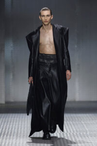 Male model walking runway in a long black leather coat open to reveal bare chest and black wide-leg pants, glossy slicked-back hair.
